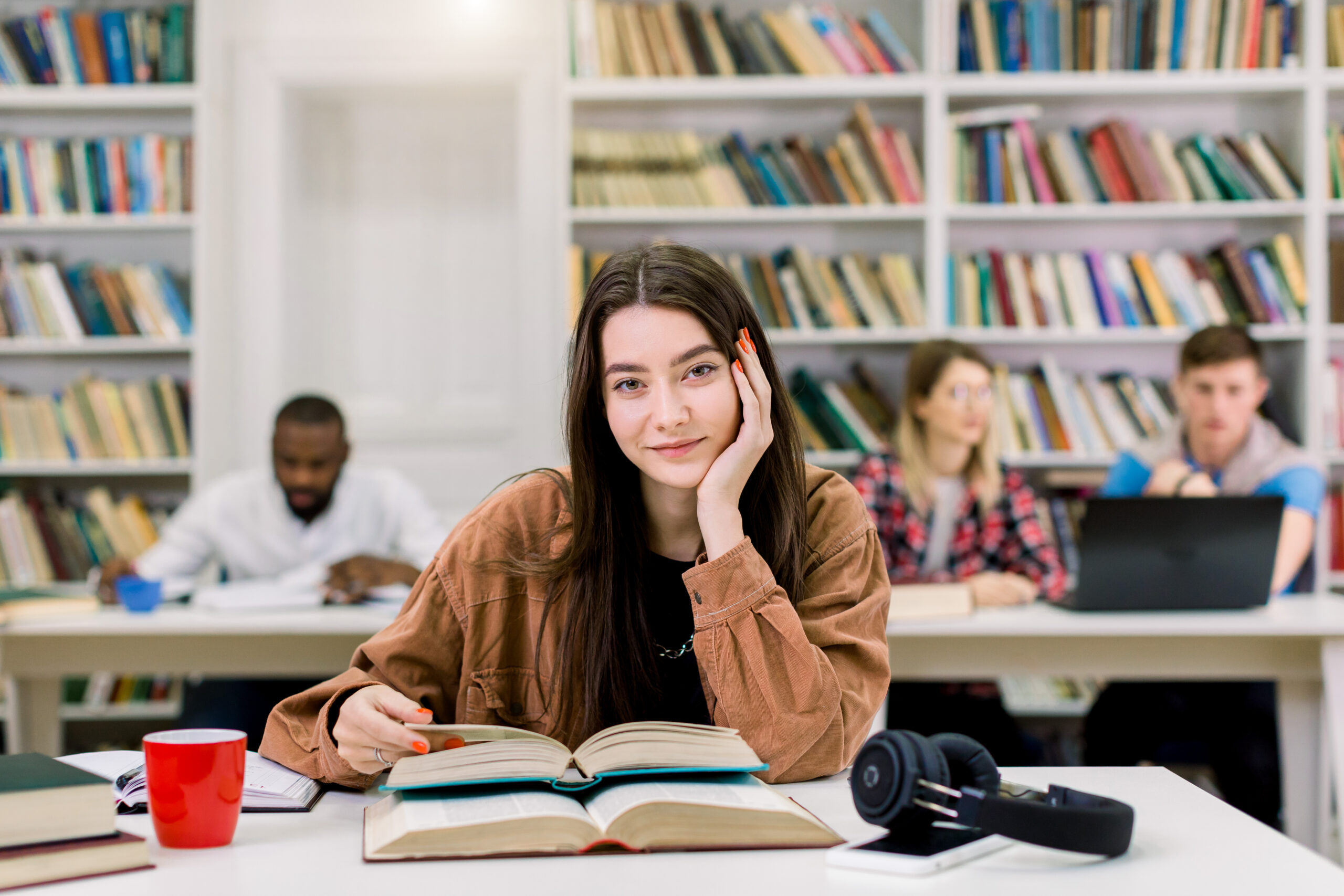 Young beautiful girl with straight long dark hair wearing in brown shirt, sitting at the table in library and preparing for test or exam, reading books. Girl posing on camera with smile Two female student friends wearing protective face masks greeting each other bumping elbows while sitting at university library, healthcare concept, precautions, new normal